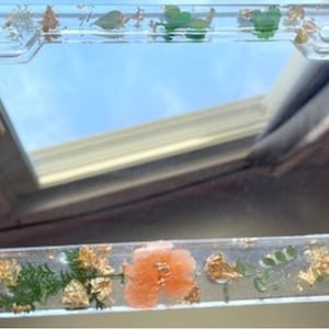 May include: A clear plastic license plate frame with a gold border and pressed flowers and leaves. The frame is decorated with gold leaf and has a clear background.