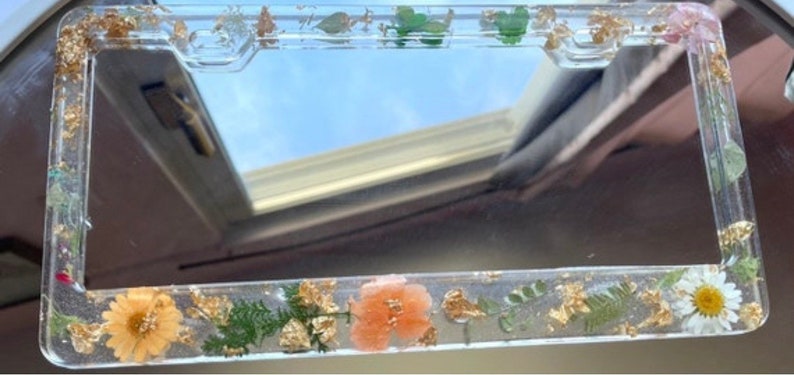 May include: A clear plastic license plate frame with a gold border and pressed flowers and leaves. The frame is decorated with gold leaf and has a clear background.