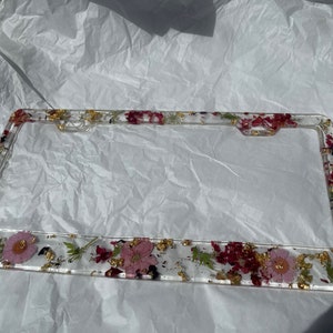May include: A clear plastic license plate frame with a floral design. The frame is decorated with pressed pink and red flowers, green leaves, and gold flakes.