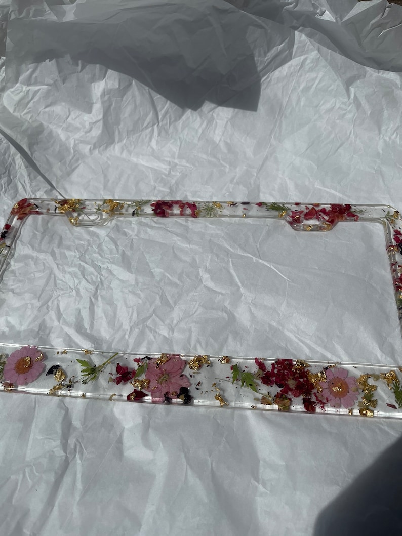 May include: A clear plastic license plate frame with a floral design. The frame is decorated with pressed pink and red flowers, green leaves, and gold flakes.