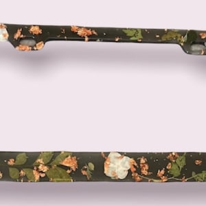 May include: Black license plate frame with a floral design featuring white and brown flowers and gold accents.