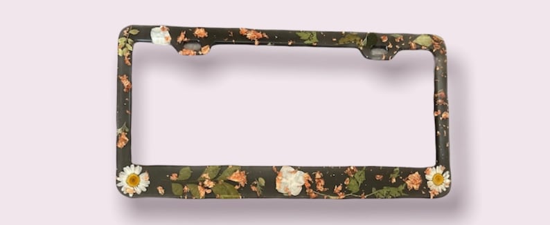 May include: Black license plate frame with a floral design featuring white and brown flowers and gold accents.