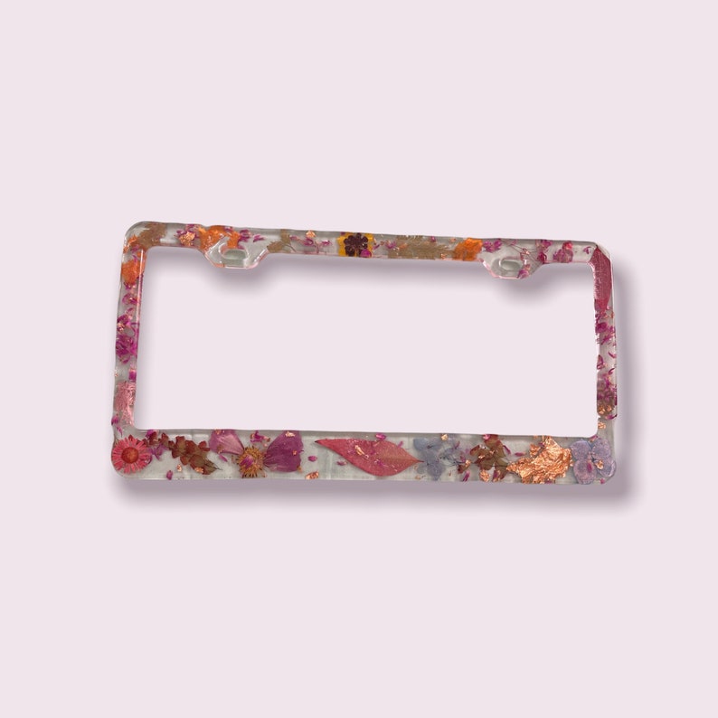 May include: A clear license plate frame with a floral design. The frame is decorated with pressed flowers and leaves in shades of pink, orange, and gold.