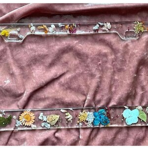 May include: A clear plastic license plate frame with pressed flowers and leaves. The frame has a light blue, yellow, white, and pink floral design.