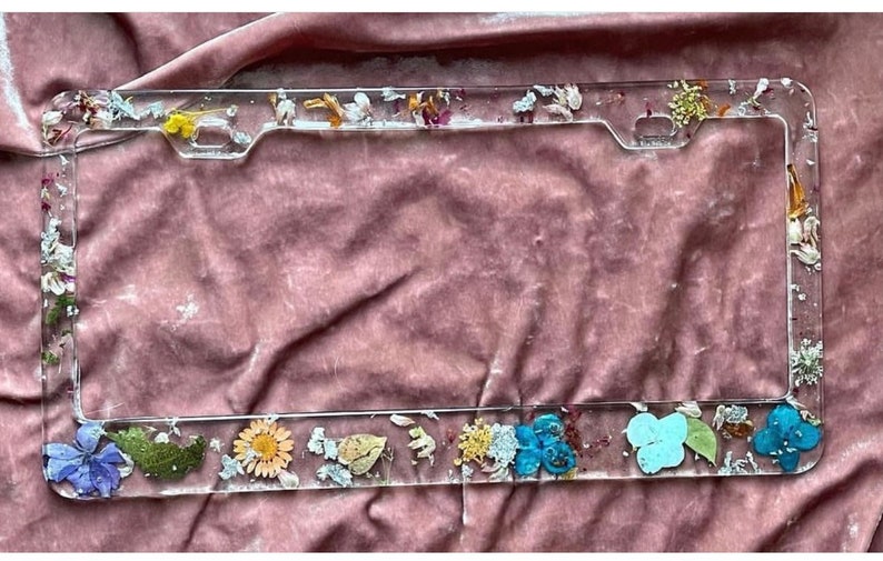 May include: A clear plastic license plate frame with pressed flowers and leaves. The frame has a light blue, yellow, white, and pink floral design.