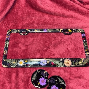 May include: Black license plate frame with a floral design featuring pressed flowers and leaves. Two matching round coasters with a similar floral design are also included.