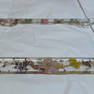 May include: A clear plastic license plate frame with a pressed flower design. The frame is decorated with dried flowers, including purple, pink, yellow, and white flowers, and gold leaf accents.
