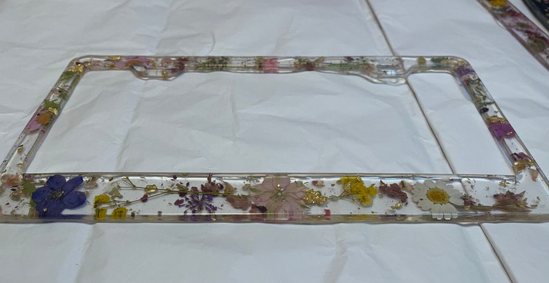 May include: A clear plastic license plate frame with a pressed flower design. The frame is decorated with dried flowers, including purple, pink, yellow, and white flowers, and gold leaf accents.