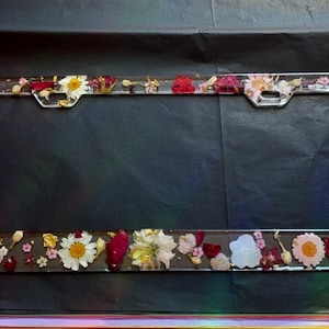 Könnte beinhalten: Ein transparenter Kennzeichenrahmen mit einem roten, weißen und goldenen Blumendesign. Der Rahmen ist aus Harz gefertigt und hat eine glänzende Oberfläche.