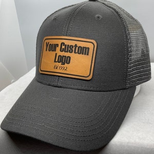 May include: A gray and black trucker hat with a brown leather patch that says "Your Custom Logo Est. 1992".