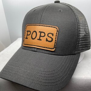 May include: A black and gray trucker hat with a brown leather patch that says "POPS" in black lettering.
