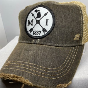 May include: A brown and beige distressed trucker hat with a black and white patch featuring the state of Michigan, the letters "MI", and the year "1837".