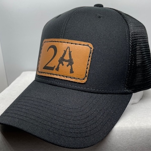 May include: Black trucker hat with a brown leather patch that says "2A" with a stylized image of two rifles.
