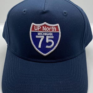 May include: A navy blue baseball cap with a white and red embroidered patch that says "UP North MICHIGAN 75".