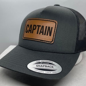May include: A gray and black trucker hat with a brown leather patch that says "CAPTAIN" on it. The hat has a black mesh back and a white sticker that says "The Authentic Yupoong Snapback Yupoong Since 1974".