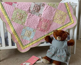 May include: A pink and green patchwork baby quilt with a scalloped edge, draped over a white crib with a teddy bear wearing a gray dress sitting in front of it.