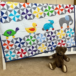 May include: A colorful baby quilt with a whimsical design featuring a turtle, duck, bunny, and elephant. The quilt is made with a variety of fabrics in shades of blue, green, yellow, red, and purple. The quilt is displayed on a white crib with a brown teddy bear sitting in front of it.