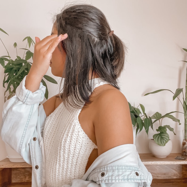 May include: A white halter top with a textured knit design. The top is paired with a light blue denim jacket. The image is set against a backdrop of green plants and a neutral wall.