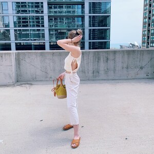 May include: A cream-colored, crocheted crop top with side ties, paired with white pants and brown woven sandals. A small, olive green handbag with a brown leather strap is held in the hand. The background features a modern building.