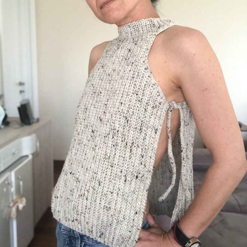 May include: A sleeveless, high-neck knit top in a speckled beige and black yarn. The sides are open with tie closures. The top is a unique, handmade garment.