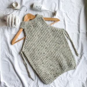 May include: A knitted sleeveless top in a speckled off-white and gray yarn, displayed on a wooden hanger. The top features a high neckline and side ties. The background includes a white surface and decorative items.