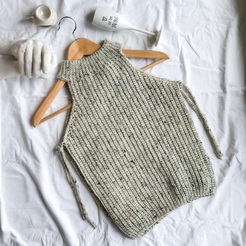 May include: A knitted sleeveless top in a speckled off-white and gray yarn, displayed on a wooden hanger. The top features a high neckline and side ties. The background includes a white surface and decorative items.