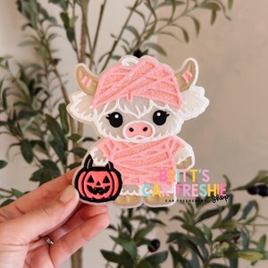 May include: A car freshie shaped like a white Highland cow dressed as a mummy in pink glittery bandages, holding a black and pink jack-o'-lantern bucket. The text BRITT'S CAR FRESHIE SHOP is printed in colorful letters on the lower right side.