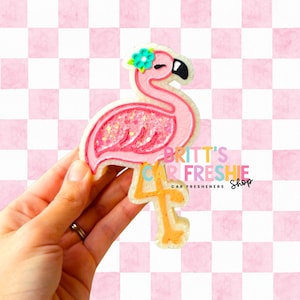 May include: A pink flamingo car air freshener with a turquoise flower and a black beak. The flamingo has a glittery body and is held against a pink and white checkered background. The text "Britt's Car Freshie Shop" is visible.