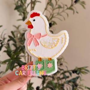 May include: A white chicken-shaped car freshie featuring a red comb, a yellow beak, and a glittery pink bow around its neck. The chicken wears green rain boots with pink flower accents. The text BRITT'S CAR FRESHIE Shop is printed on the front.