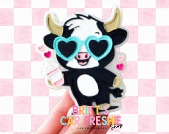 May include: A black and white cow-shaped car air freshener featuring light blue heart-shaped sunglasses and a pink coffee cup. The cow has tan horns and is surrounded by small pink hearts. The text BRITT'S CAR FRESHIE SHOP is visible.