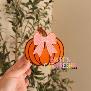 May include: An orange pumpkin-shaped car freshie featuring a textured surface, a dark brown stem, and a light pink glittery bow attached to the front. The text BRITT'S CAR FRESHIE Shop is printed in colorful letters on the right side of the pumpkin.