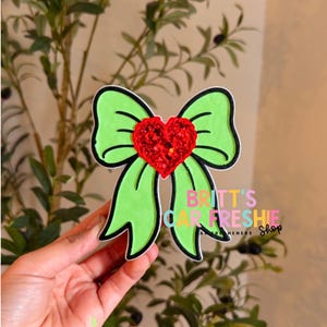 May include: A lime green bow-shaped air freshener featuring a central red heart covered in red glitter. The bow has a thick black outline and the text BRITT'S CAR FRESHIE SHOP is printed in colorful letters on the lower right side of the bow.