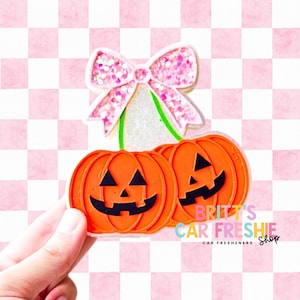 May include: A car air freshener shaped like two orange jack-o'-lantern pumpkins with black facial features, connected by green stems and topped with a large, glittery pink bow. The text BRITT'S CAR FRESHIE SHOP is printed on the right side.