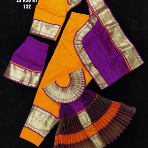 Puede incluir: Un conjunto de traje de danza tradicional indio en naranja y morado. El conjunto incluye una blusa, una falda y una pieza plisada en forma de abanico. La tela tiene ribetes dorados y un patr&oacute;n de rayas. El texto en la imagen dice "BH SUN SHP DVNY 29 K30 K7 132".