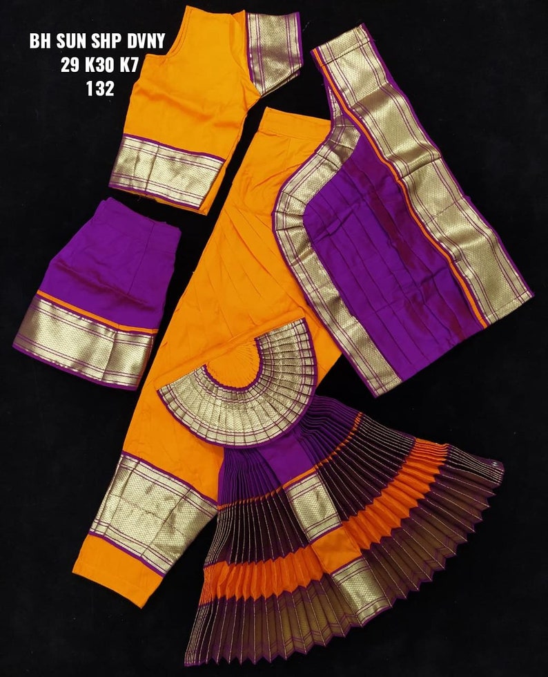 Puede incluir: Un conjunto de traje de danza tradicional indio en naranja y morado. El conjunto incluye una blusa, una falda y una pieza plisada en forma de abanico. La tela tiene ribetes dorados y un patr&oacute;n de rayas. El texto en la imagen dice "BH SUN SHP DVNY 29 K30 K7 132".
