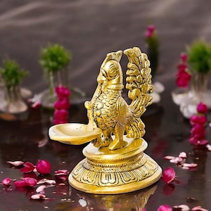 May include: A gold metal peacock diya, a traditional Indian oil lamp, with intricate details. The diya is on a round base with a decorative design.