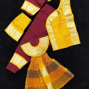 Puede incluir: Un conjunto de traje de danza tradicional indio en color marr&oacute;n y amarillo. El conjunto incluye una falda larga con pliegues, una blusa de manga corta, una parte superior a juego y una pieza decorativa para la cabeza. La tela est&aacute; adornada con ribetes dorados.