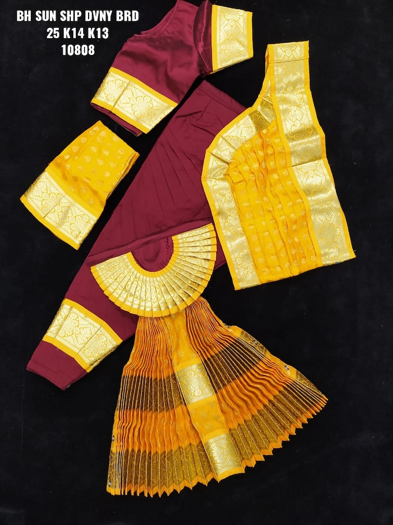 Puede incluir: Un conjunto de traje de danza tradicional indio en color marr&oacute;n y amarillo. El conjunto incluye una falda larga con pliegues, una blusa de manga corta, una parte superior a juego y una pieza decorativa para la cabeza. La tela est&aacute; adornada con ribetes dorados.