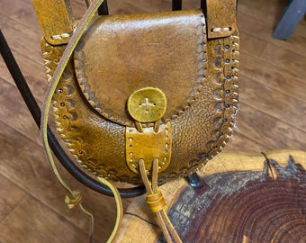May include: A small brown leather bag with a flap closure and a decorative button. The bag has a long strap and is hanging from a wooden chair.