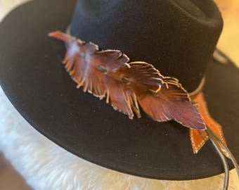 May include: A black fedora-style hat with a decorative leather feather accent. The feather is a mix of brown and orange hues, and the hat has a wide brim. A thin leather cord wraps around the base of the hat.