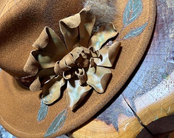 May include: A brown fedora hat with a decorative leather flower and feather accents. The hat features painted leaf designs in teal and brown. The brim is slightly curved, and the hat is resting on a wooden surface.