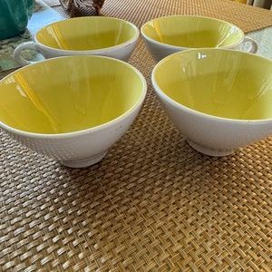 May include: Four white teacups with yellow interiors. The teacups are arranged on a woven brown placemat.