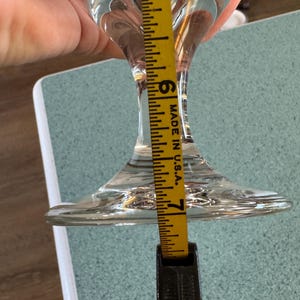 May include: A clear glass stemmed wine glass with a flared base. A yellow measuring tape shows the glass is approximately 7 inches tall. The tape is marked "MADE IN U.S.A."