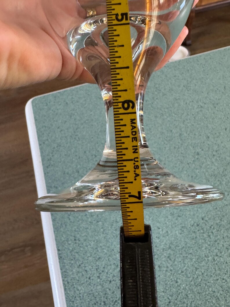 May include: A clear glass stemmed wine glass with a flared base. A yellow measuring tape shows the glass is approximately 7 inches tall. The tape is marked "MADE IN U.S.A."