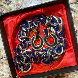May include: A blue and gold chain necklace and matching earrings in a red velvet box. The necklace is made of linked circles with a gold trim. The earrings are hoop style with a blue enamel center.