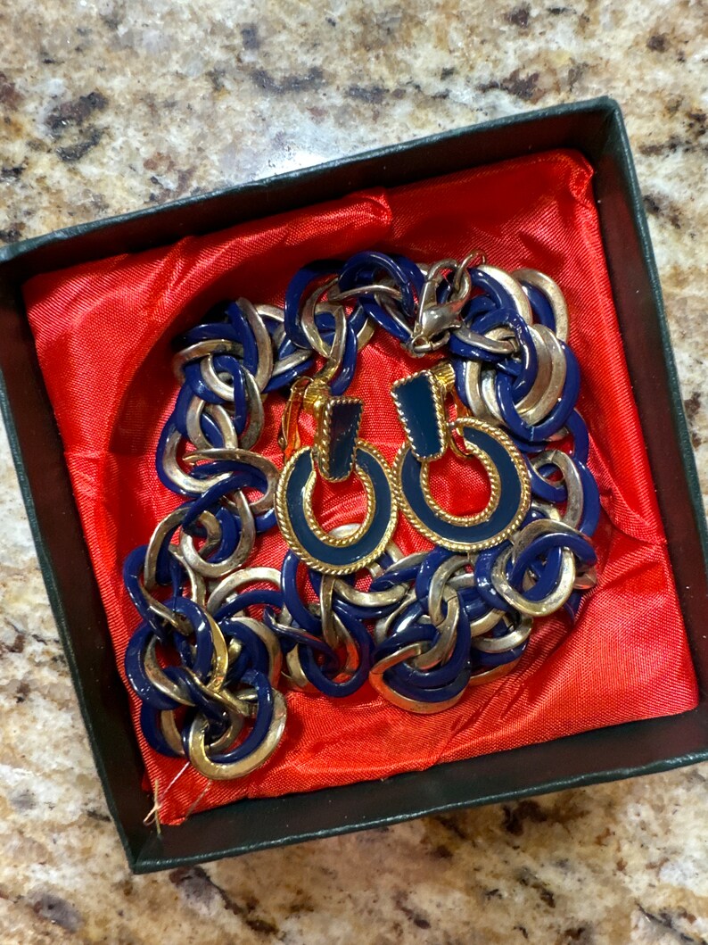 May include: A blue and gold chain necklace and matching earrings in a red velvet box. The necklace is made of linked circles with a gold trim. The earrings are hoop style with a blue enamel center.