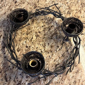 May include: A black metal wreath with three twig nests for candles. The wreath is made of twisted branches and has a rustic look.