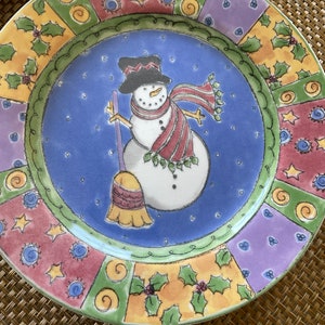 May include: A colorful ceramic plate with a whimsical snowman design. The plate features a patchwork of green, yellow, red, purple, and blue with a white snowman holding a broom in the center.