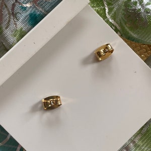May include: A pair of gold-toned earring backs. The backs are made of metal and have a small, clear stone in the center.