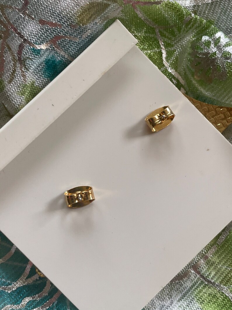 May include: A pair of gold-toned earring backs. The backs are made of metal and have a small, clear stone in the center.